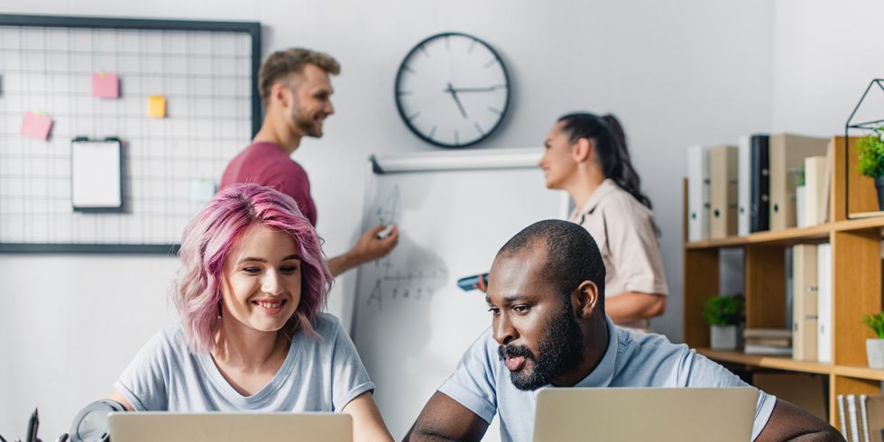 Zwei Personen sitzen im Büro vor ihren Laptops, im Hintergrund stehen zwei Menschen vor einem Flipchart